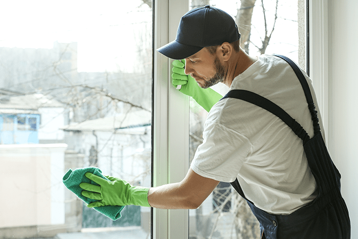 Man in a white t-shirt and black cap cleaning a window with a green cloth.
